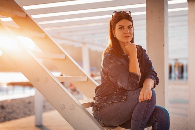 Una joven y hermosa mujer segura de sí misma posa sentada en una escalera de madera blanca puesta de sol en el fondo