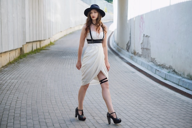 Joven hermosa mujer sexy vistiendo ropa de moda, vestido blanco, sombrero negro posando en la calle de la ciudad
