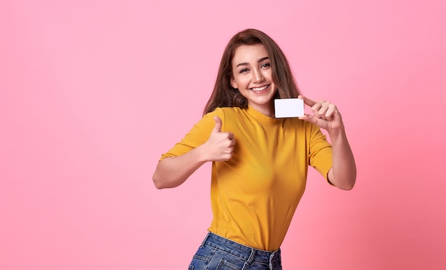 Joven hermosa mujer sonriente en camisa amarilla mostrando tarjeta de crédito en mano sobre fondo rosa