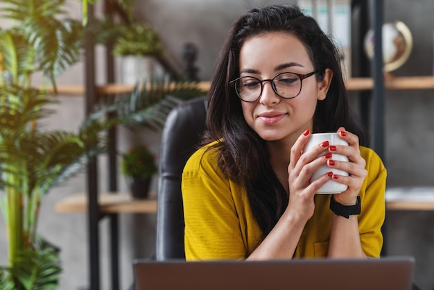 Joven hermosa mujer sonriente disfrutando de su taza de café mientras trabaja o estudia en una laptop