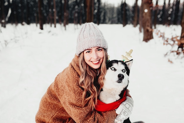 Joven hermosa mujer sonriente feliz en abrigo de piel abrazando perro husky vestida con bufanda roja de Navidad y cuernos de ciervo festivo. Foto de retrato de niña y perro en la nieve blanca en el bosque de invierno. Feliz Año Nuevo