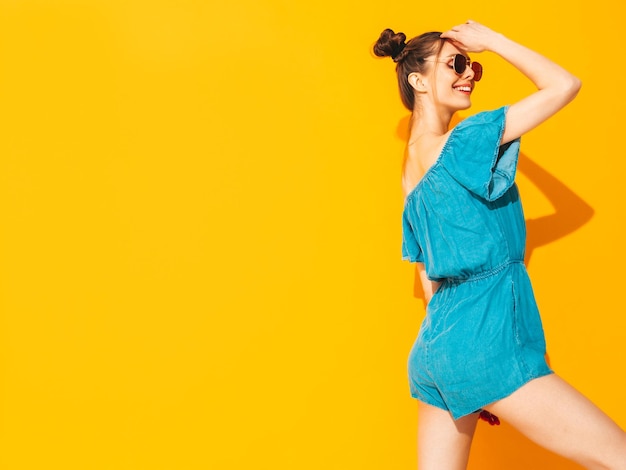 Joven hermosa mujer sonriente en overoles de jeans de verano de moda Mujer sexy despreocupada con peinado de dos cuernos posando junto a la pared amarilla en el estudio Modelo positivo divirtiéndose Aislado en gafas de sol