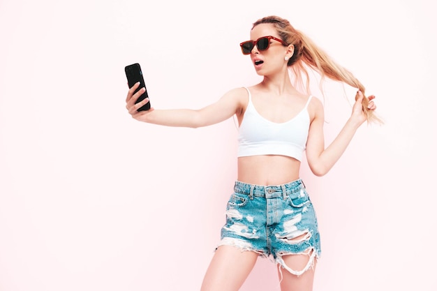 Joven hermosa mujer sonriente en pantalones vaqueros de moda de verano Mujer rubia despreocupada sexy posando cerca de la pared en el estudio Modelo positivo divirtiéndose Alegre y feliz en gafas de sol Aislado Tomando selfie