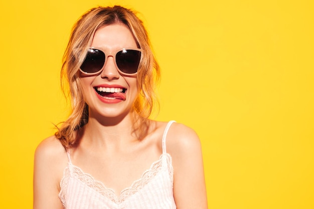 Joven hermosa mujer sonriente en ropa de verano de moda Mujer sexy despreocupada posando cerca de azul amarillo en el estudio Modelo positivo divirtiéndose Alegre y feliz En gafas de sol Muestra lengua