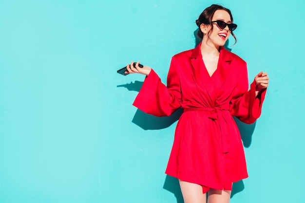 Foto joven hermosa mujer sonriente en vestido rojo de verano de moda mujer despreocupada sexy posando junto a la pared azul en el estudio modelo positivo divirtiéndose alegre y feliz en gafas de sol