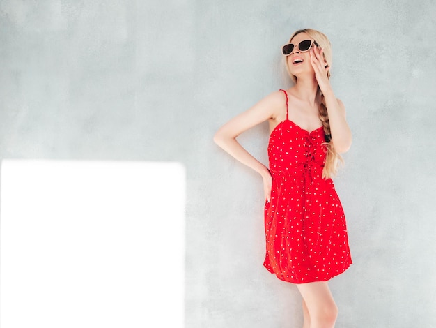 Joven hermosa mujer sonriente en vestido rojo de verano de moda Sexy mujer rubia despreocupada posando cerca de la pared en el estudio Modelo positivo divirtiéndose Alegre y feliz en gafas de sol En un día soleado