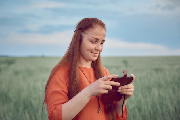 Joven hermosa mujer vestida de rojo con el pelo rojo mira tus fotos en tu teléfono y se para en un campo de trigo verde por la noche al atardecer tecnología digital moderna