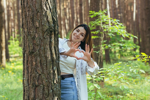 Joven hermosa mujer voluntaria en el bosque cerca de un árbol sonriendo mira a la cámara y muestra las manos y los dedos un corazón un signo de amor por la naturaleza