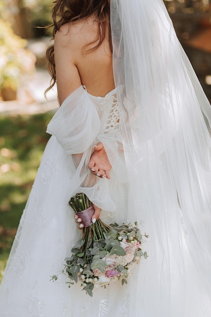 joven y hermosa novia con cabello largo y marrón en un vestido de novia al aire libre con un ramo de flores de boda retrato en toda su longitud