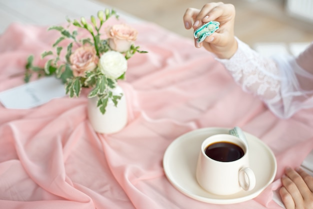 Joven hermosa novia caucásica disfrutando del desayuno de macarrones franceses y café en una mesa de madera con un mantel de gasa rosa y un jarrón de flores
