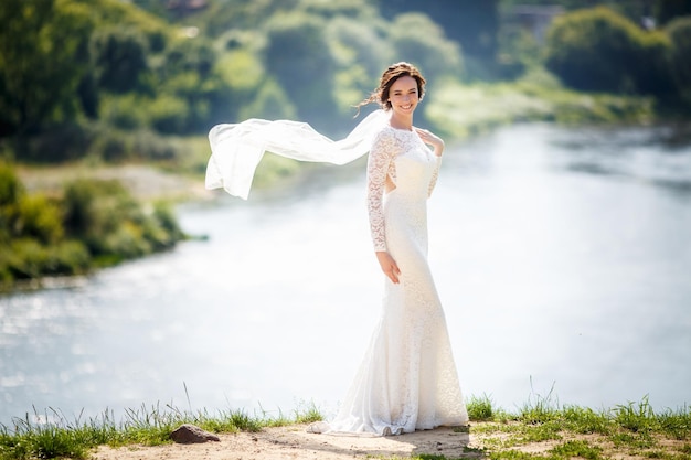 Joven hermosa novia se encuentra cerca del río, el viento sopla el velo