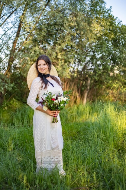 Una joven y hermosa novia con maquillaje y peinado se para en un prado con un vestido de novia estilo boho con un ramo de flores en las manos, un gran sombrero de paja en el hombro