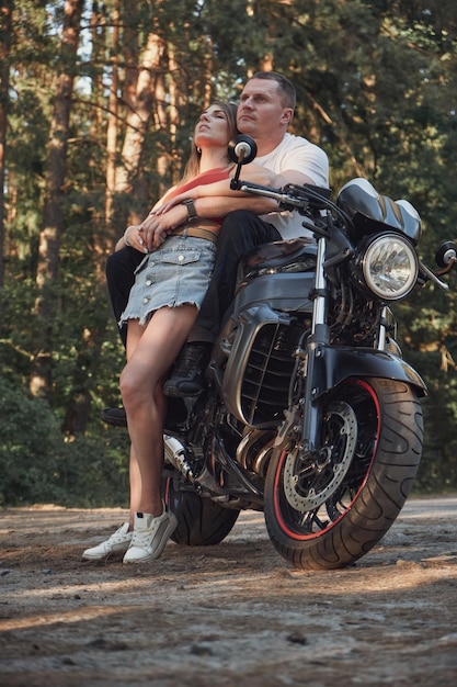 Una joven y hermosa pareja abrazándose sentada en una motocicleta viajan juntos por un camino forestal