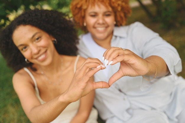 Joven hermosa pareja afroamericana lesbiana feliz sentada sobre la hierba verde mostrando signo de amor