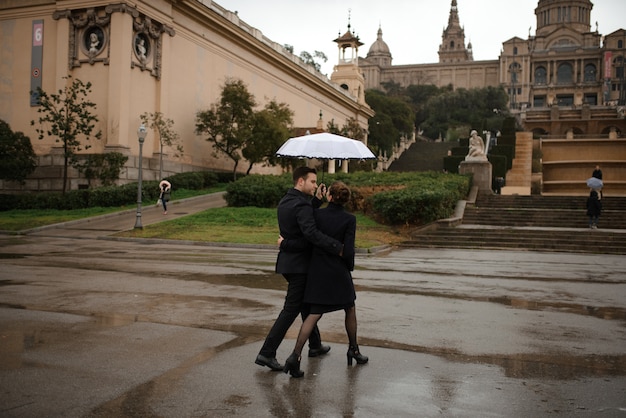 Joven hermosa pareja amorosa hispana camina bajo un paraguas durante la lluvia en la Plaza España. Pareja posando en el contexto del Museo Nacional de Arte de Cataluña.