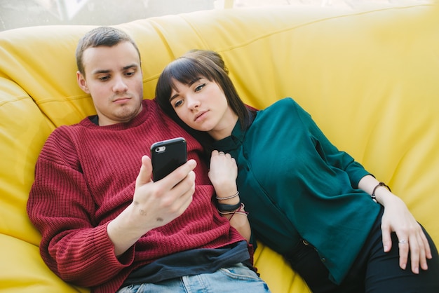 Joven hermosa pareja de enamorados tumbado en una silla amarilla y utiliza un teléfono móvil en su tiempo libre.