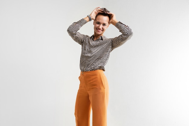 Una joven hermosa con el pelo corto y oscuro, maquillaje con una camisa a rayas blancas y negras, pantalones marrones y zapatos, se levanta y sonríe.