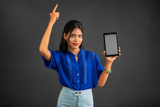 Una joven hermosa que muestra una pantalla en blanco de un teléfono inteligente o un teléfono móvil o una tableta en un fondo gris