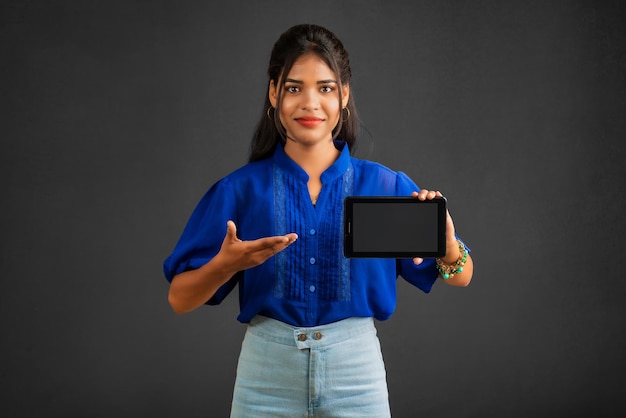 Una joven hermosa que muestra una pantalla en blanco de un teléfono inteligente o un teléfono móvil o una tableta en un fondo gris