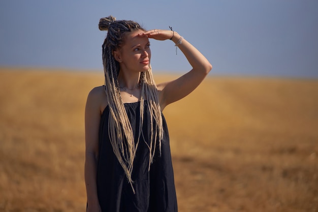 Una joven hermosa con rastas se encuentra en medio de un campo con pasto seco y mira ...