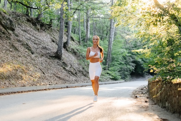 Una joven hermosa en ropa deportiva blanca está corriendo, en la carretera en un denso bosque, durante la puesta de sol. Hacer deporte al aire libre. Un estilo de vida saludable.