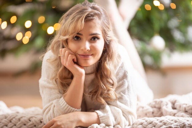 Joven hermosa rubia sonriente acostada sobre una manta junto a un árbol de Navidad