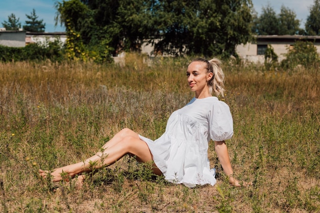 Una joven y hermosa rubia con un vestido blanco se sienta en el césped sin zapatos