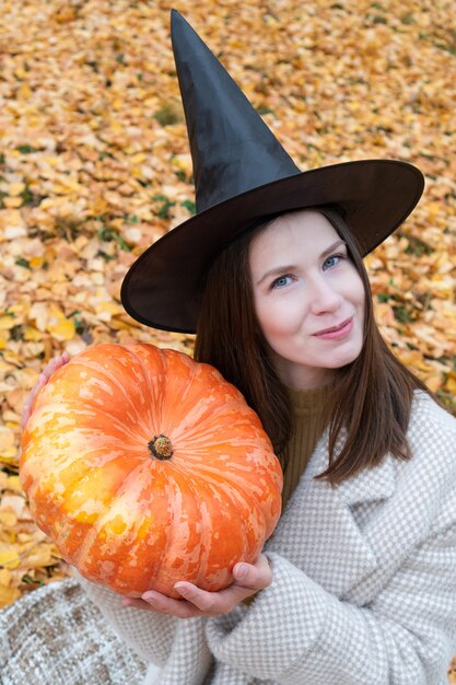 Una joven hermosa con un sombrero de bruja y un elegante abrigo sostiene una gran calabaza en sus manos