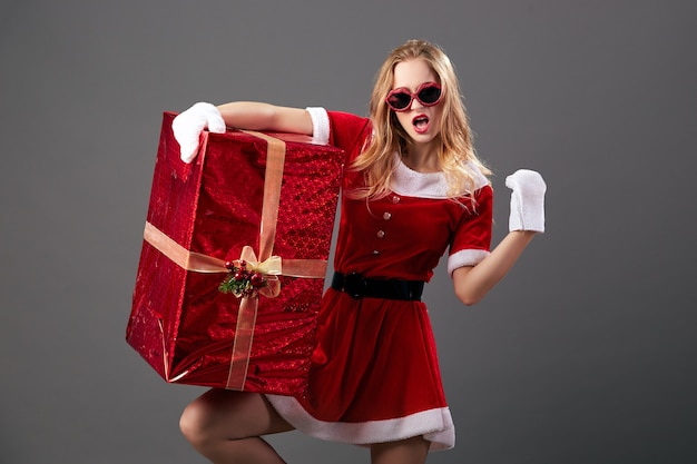 La joven y hermosa Sra. Santa Claus con gafas de sol vestida con la túnica roja, guantes blancos y tacones altos sostiene el enorme regalo de Navidad sobre el fondo gris. .