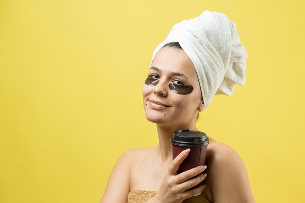 Una joven hermosa con una toalla blanca en la cabeza usa parches de gel de colágeno debajo de los ojos Máscara debajo de la cara de tratamiento de los ojos La niña sostiene un vaso de café en las manos