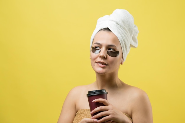 Una joven hermosa con una toalla blanca en la cabeza usa parches de gel de colágeno debajo de los ojos Máscara debajo de la cara de tratamiento de los ojos La niña sostiene un vaso de café en las manos