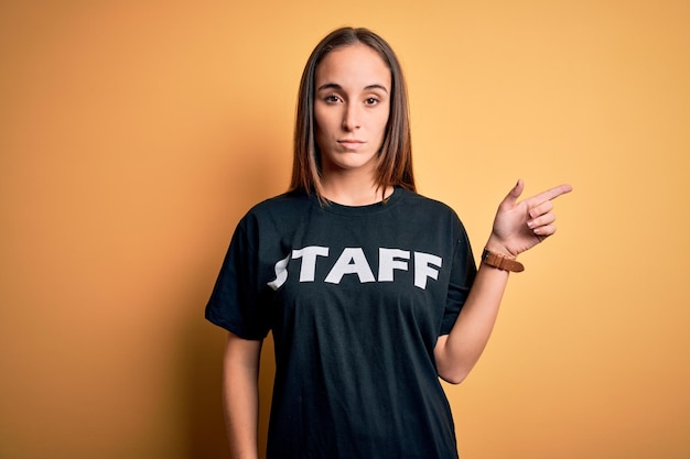 Joven y hermosa trabajadora con camiseta de uniforme de personal sobre fondo amarillo aislado Señalando con el dedo de la mano hacia un lado mostrando publicidad cara seria y tranquila