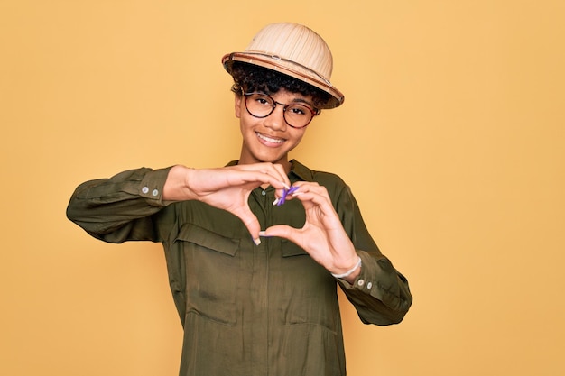 Joven hermosa turista afroamericana con gafas y sombrero de explorador sonriendo enamorada haciendo forma de símbolo de corazón con las manos Concepto romántico