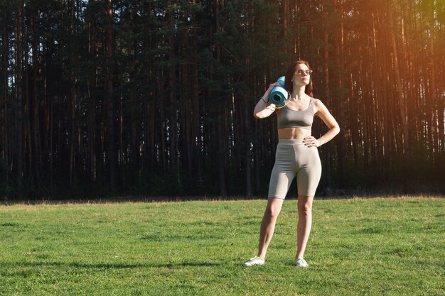 Una joven hermosa con uniforme deportivo y una estera de yoga en sus manos vino a practicar deportes. Entrenamiento al aire libre, en el parque, verano, soleado. Yoga en el concepto de parque.