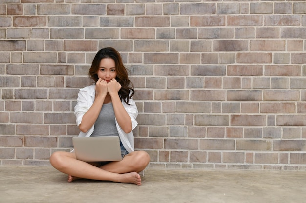 Una joven hermosa usando una computadora portátil mientras está sentada contra la pared en casa