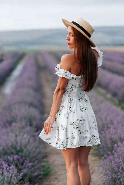 Una joven hermosa con un vestido y un sombrero delicados camina por un hermoso campo de lavanda y disfruta de la fragancia de las flores Descanso y hermosa naturaleza Floración de lavanda y recolección de flores