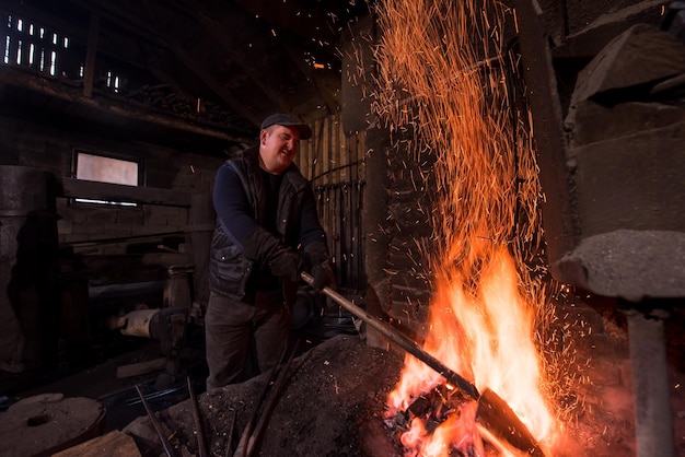 joven herrero tradicional trabajando con fuego abierto El herrero haciendo llamas en la herrería con chispas de fuegos artificiales forjando hierro caliente en el taller