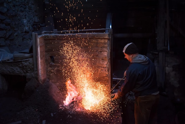 joven herrero tradicional trabajando con fuego abierto El herrero haciendo llamas en la herrería con chispas de fuegos artificiales forjando hierro caliente en el taller