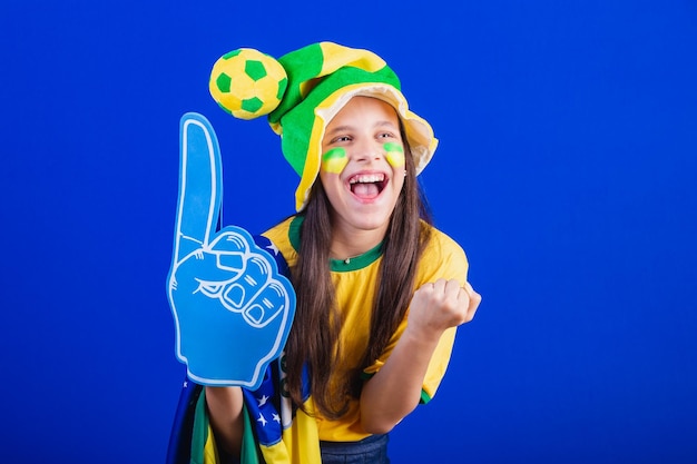 Joven hincha de fútbol de Brasil vestida con sombrero y bandera usando un dedo de espuma festejando increíble