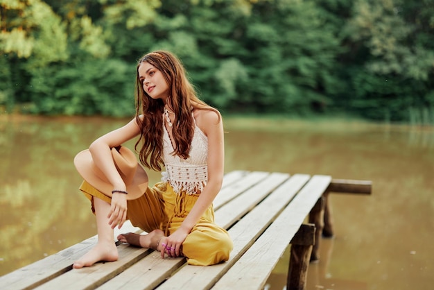 Una joven hippie se sienta en un puente del lago con ropa ecológica elegante y sonriendo