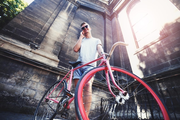 Joven hipster atractivo con camiseta gris y pantalones cortos de mezclilla azul con gafas de sol que se quedan en bicicleta y hablan por un teléfono móvil en la calle de la ciudad de fondo