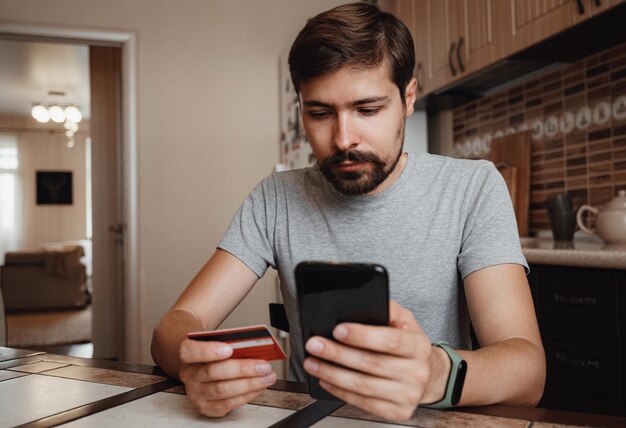 Joven hipster comprando en línea con tarjeta de crédito usando un teléfono inteligente en casa