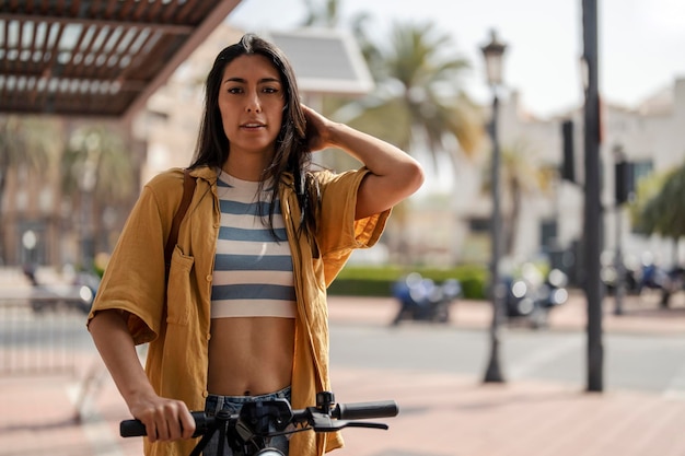 Una joven hipster está parada en la calle de la ciudad en una ciudad de Europa con su scooter ecológico