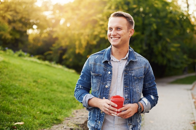 Joven Hipster hombre de pie con café para llevar en el parque, sonriendo complacientemente a la cámara. Feliz chico guapo despreocupado en chaqueta vaquera azul