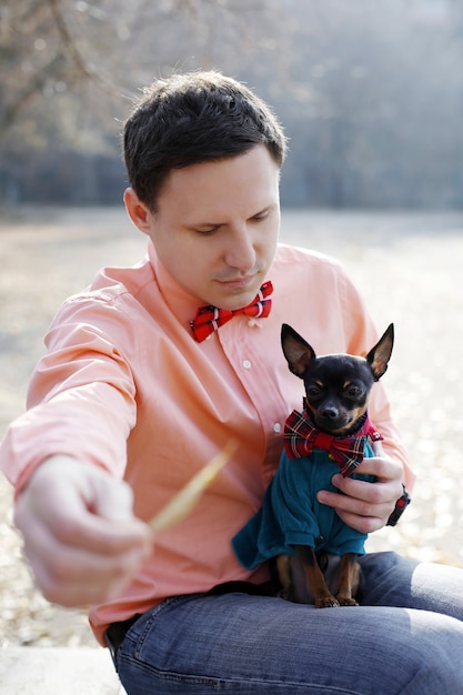Joven hipster jugar con perro terrier de juguete al aire libre, mirada familiar con lazo de corbata