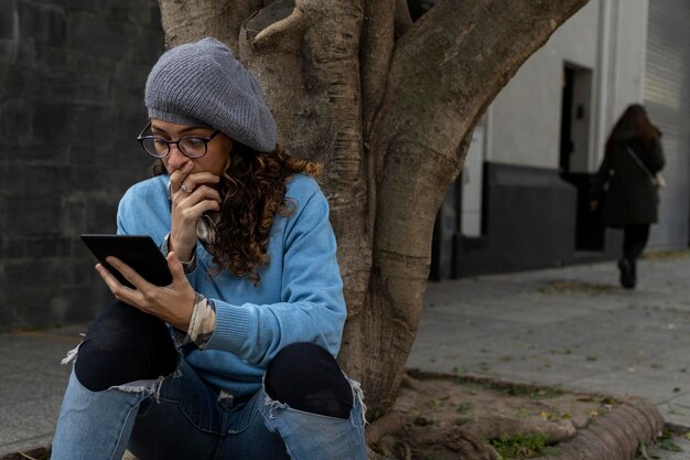 Joven hipster latinoamericana sentada en la acera leyendo un libro digital