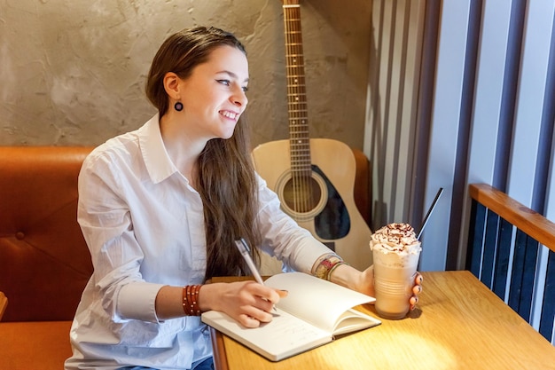 Joven hipster sonriente sentada tocando la guitarra y escribiendo una canción en el café. Chica aprendiendo a tocar música