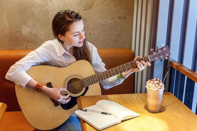 Joven hipster sonriente sentada tocando la guitarra y escribiendo una canción en el café. Chica aprendiendo a tocar música