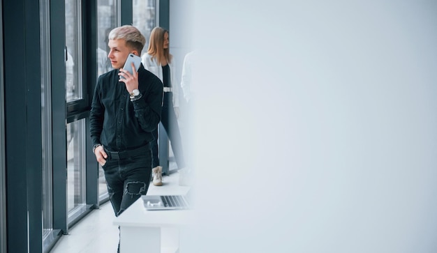 Joven hipster con teléfono parado frente a un grupo de jóvenes exitosos que trabajan y se comunican juntos en el interior de la oficina