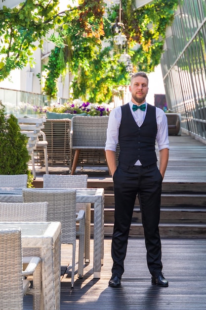 Un joven hipster con traje y corbata de moño, un gerente en un café callejero de verano espera a los visitantes antes de abrir